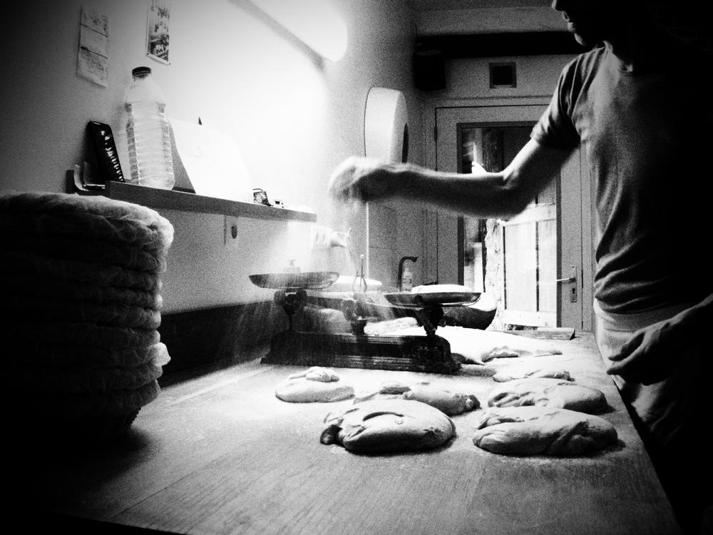 photo noir & blanc d'un boulanger en train de fleurer des pâtons (saupoudrer de farine), sur un plan de travail en bois.